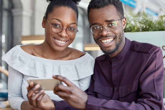 Two dark skinned woman and man with positive smiles, watch video on smart phone, wear glasses, dressed in fashionable clothes, have pleasant expressions. People, ethnicity and technology concept.