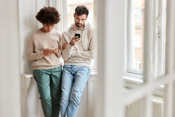 Youngsters of different races use modern gadgets for surfing internet, ignore real communication, read news on webpage, stand near window sill in spacious room, show their addiction to phones