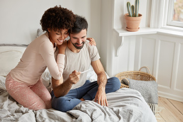 Pregnancy news and family concept. Cheerful dark skinned woman embraces her husband, shows positive...
