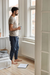 Indoor full length shot of thoughtful unshaven man in casual t shirt and trousers, stands near...
