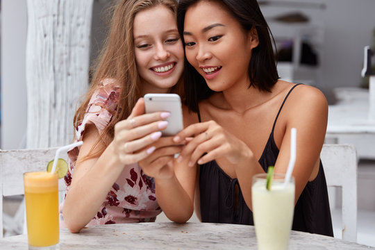 Cropped shot of multiethic young women watch online broadcasting on cell phone, search information in internet, drink fresh cocktails in restaurant, enjoy spare time, sit indoor. Leisure and lifestyle