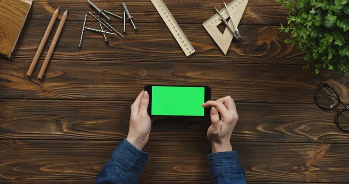Top view on the black smartphone lying horizontally on the wooden table with office supplies and nails, female hands scrolling and tapping on the green screen. Chroma key.