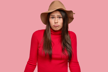 Fototapeta premium Uncertain brunette woman has clueless expression, looks in disbelief, purses lips, wears stylish brown hat, red sweater, poses against pink studio wall. Puzzled student cant find answer during exam