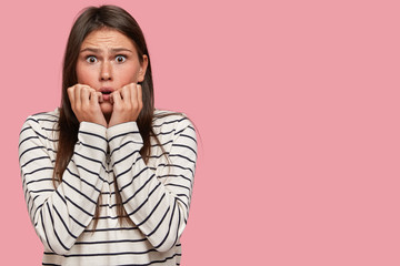 Frightened emotive European woman keeps both hands near mouth, stares with eyes popped out, bites...