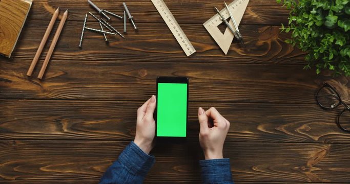 Top view on the black smartphone lying vertically on the wooden table with office supplies and nails, female hands scrolling and tapping on the green screen. Chroma key.