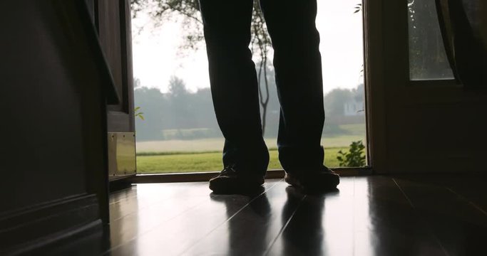 A door is opened and a man back lit or silhouetted by the daylight outdoors stands momentarily in the doorway then walks calmly inside.