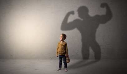 Little waggish boy in an empty room with musclemen shadow behind

