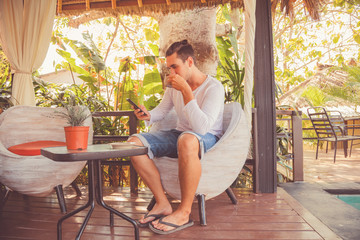 Attractive man using cellphone and drinking coffee outdoors.