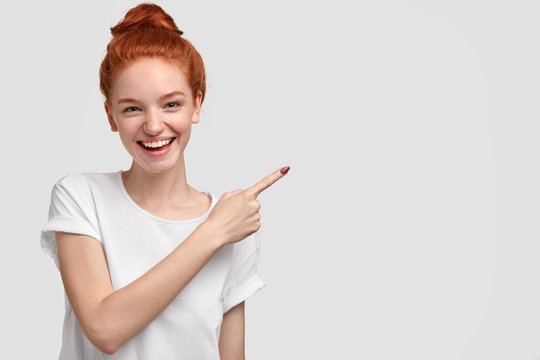 Glad satisfied red haired young female with charming smile, shows free space for your promotional content, advertises something against white background, dressed casually. Look aside, please.
