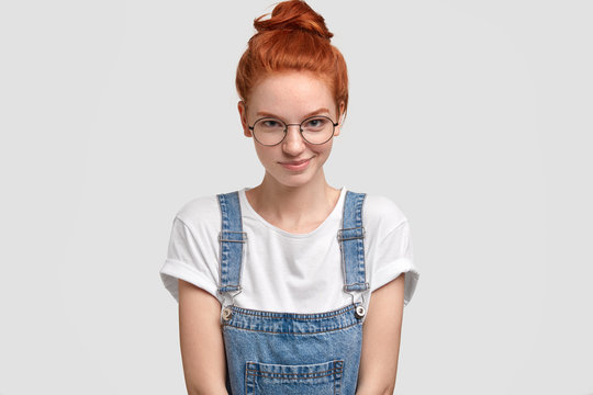 Horizontal shot of pleasant looking ginger young European woman in round transparent glasses, stylish dungarees, poses against white background, has glad expression. People and emotions concept.