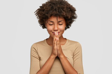 Meditation concept. Beautiful young black woman stands in meditative pose, enjoys peaceful atmosphere, holds hands in praying gesture, isolated over white background, has sense of inner peace