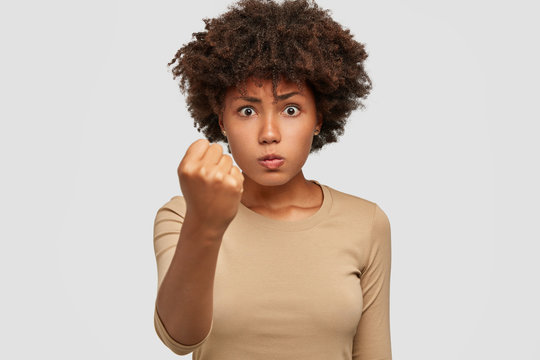 Photo of strict serious young woman dominates, shows fist at camera, threatens or warns about something, feels angry with somebody, models against white studio wall. Authority, leadership concept