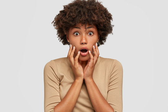 Headshot of shocked beautiful black woman opens mouth widely, keeps both hands on cheeks, cant believe in exciting news, stares with bugged eyes, dressed in beige sweater, isolated on white wall