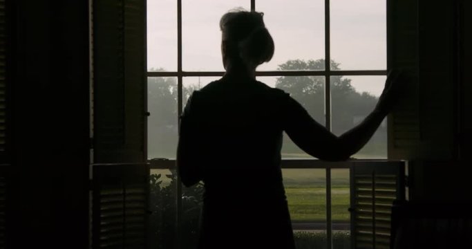 A woman silhouetted by the daylight walks to a window looking out into suburbia and drinks her coffee on a leisurely weekend morning.