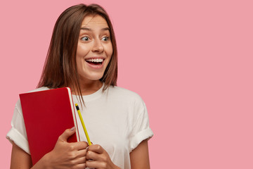 Sideways shot of glad amazed student happy to see groupmate into distance, has toothy smile, carries notebook and pencil models against pink background with copy space on right side for your promotion
