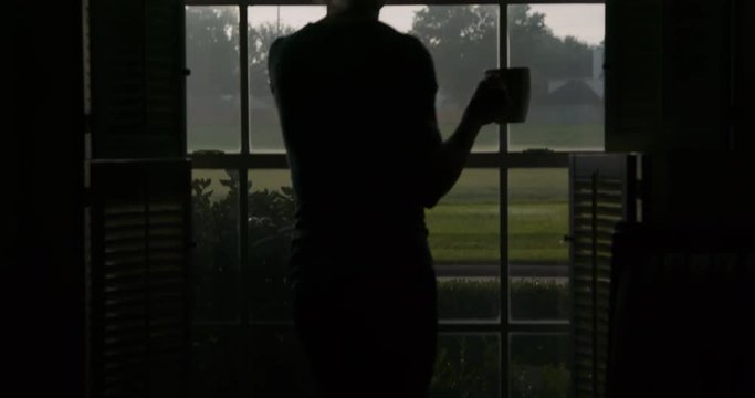 Scene moves up a woman standing by a window looking out into suburbia drinking coffee on a leisurely weekend morning who is silhouetted by the daylight outdoors.