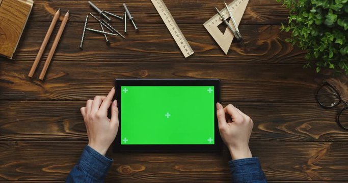 Top view on the black tablet device lying horizontally on the wooden table with office supplies and nails, female hands scrolling and tapping on the green screen. Chroma key. Tracking motion.