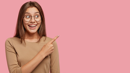 Studio shot of good looking satisfied brunette woman with toothy broad smile, wears round spectacles, points with index finger aside, shows copy space for your advertising content, being in good mood