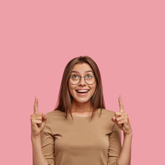 Vertical shot of good looking brunette woman with broad smile, looks joyfully upside, indicates...