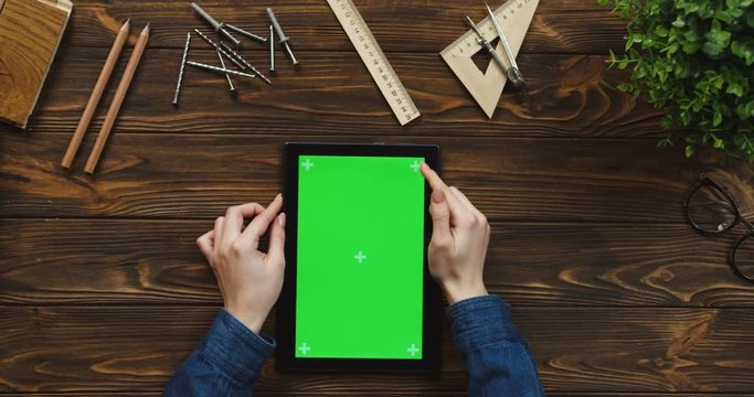 Top view on the black tablet device lying vertically on the wooden table with office supplies and nails, female hands scrolling and tapping on the green screen. Chroma key. Tracking motion.
