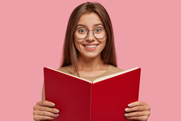 Studio shot of pleased cheerful woman has peaceful calm happy expression, holds opened textbook in front, wears optical glasses for good vision, learns material, satisfied with good knowledge