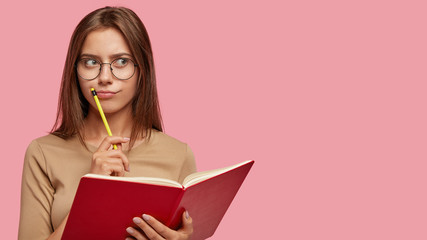 Pensive female writer ponders on new ideas, holds red notebook and pencil near mouth, has...