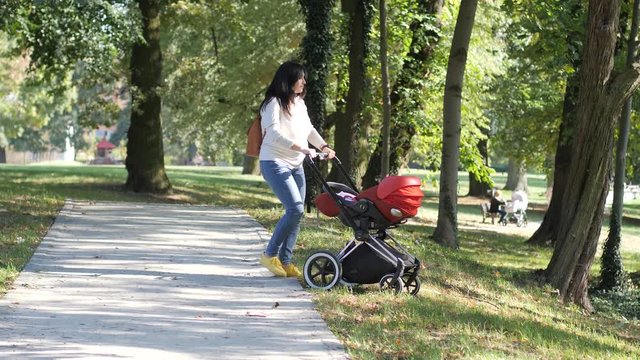 Young mother with newborn infant baby in stroller walking in city park slow motion