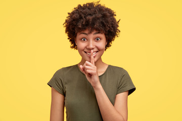 Shh, be quiet, keep voice down. Isolated shot of cheerful young African American female makes silence sign, dressed in casual t shirt, isolated over yellow background, gossips about something