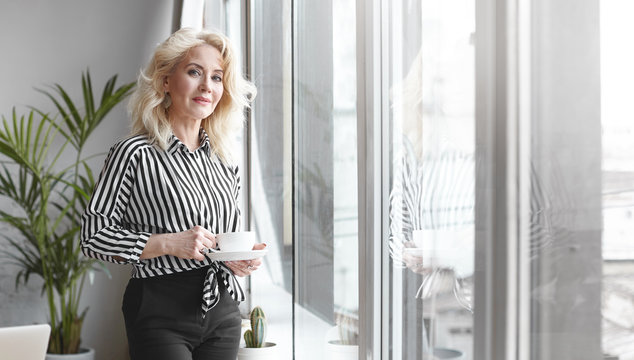 Serious attractive sixty year old businesswoman working in her modern office, standing by the window, enjoying fresh morning coffee, looking at camera with confident smile, copyspace for text