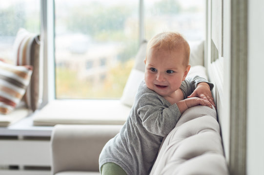 Baby Boy Crawling And Climbing On Sofa Against Big Window