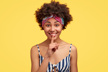 Beautiful dark skinned cheerful model wears casual striped top, colourful headband on forehead,...