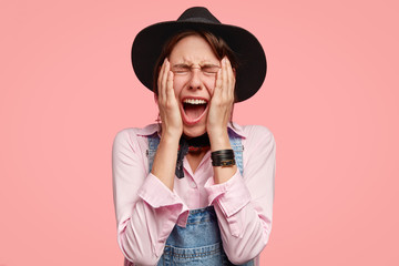 Desperate Caucasian young woman in headgear keeps both hands on cheeks, exclaims with negative...