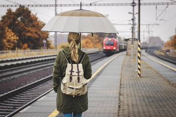 Young woman waiting for train on railway station. Backpacker with umbrella is traveling in rain at...