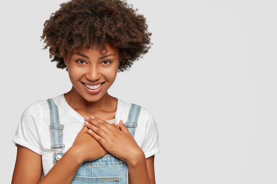 Delighted black teenage girl has toothy smile, keeps both hands on chest, being pleased or moved by heart piercing story, has Afro dark curly hair, poses over white background with copy space for logo