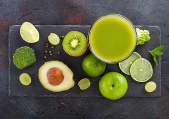 Glass of fresh raw organic green toned fruit and vegetables on stone board black background. Avocado, lime, apple, kiwi and grapes with broccoli and cauliflower