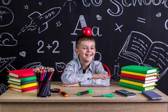 Cheerful and happy hellion schoolboy sitting at the desk with books, school supplies, with a red apple on the top of his head