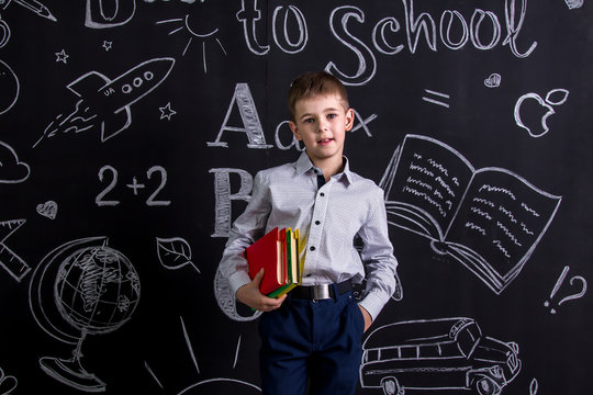 Cute schoolboy standing before the chalkboard as a background with a couple of books hugging them under the armpit. Landscape picture