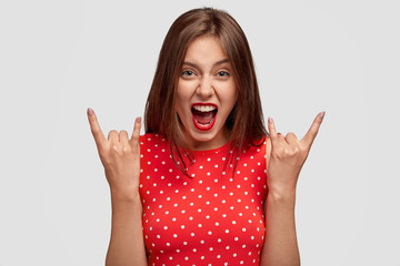 Studio view of cool European woman makes rock gesture, exclaims with madness, keeps mouth widely...
