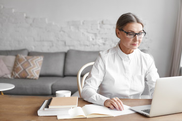 Busy modern mature female wearing spectacle and formal shirt working distantly using free wifi on...