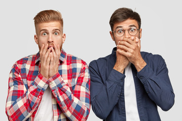 Stunned shocked two guys gasp from fear, cover mouthes with both hands, being astonished with terrible event, stand next to each other, have stylish haircuts, isolated on white blank studio wall