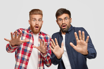 Photo of two men have scared expressions, make frightened gesture with plams, try to defend from something bad, exclaim with fear, ask not come close, stand against white background. Negative feelings