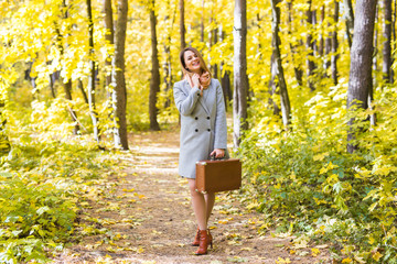 Autumn, fashion, people concept - woman with brown retro suitcase walking through the autumn park and smiling