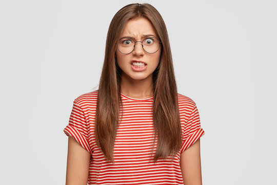 Photo of displeased Caucasian woman clenches teeth and frowns face with irritation, wears round glasses and casual striped t shirt, poses against white background. Negative emotions concept.