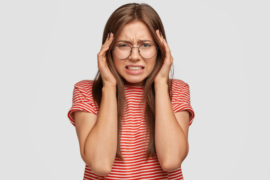 Young desperate European woman has bad headache, clenches teeth, keeps hands on head, has miserable facial expression, feels overworked, poses against white background. Omg, stop this please