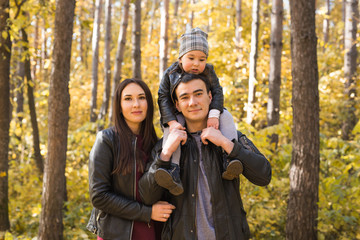 Fototapeta premium Family, autumn, people concept - young family walking in autumn park. Daughter sitting on dad's shoulders