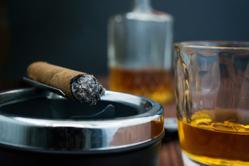 Photo of a whiskey glass close-up and a smoldering cigar on an ashtray and a blurred bottle with...