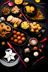 Diwali sweets and snacks arranged in a group with Diya or oil lamp, flowers and Fire Crackers or Patakhe over moody background, selective focus