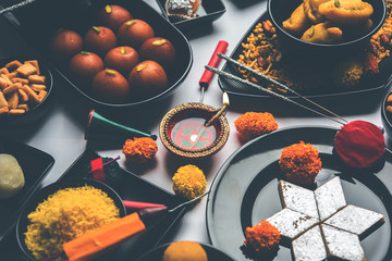 Diwali sweets and snacks arranged in a group with Diya or oil lamp, flowers and Fire Crackers or Patakhe over moody background, selective focus
