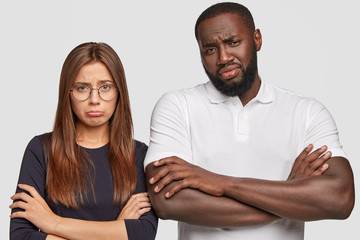 Fototapeta premium Horizontal shot of displeased young multiethnic couple have miserable looks as draw distrubing picture in imagination, keep arms folded, doesnt like something, isolated over white background