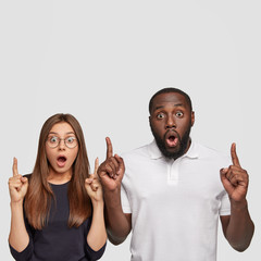 Studio shot of stupefied emotive mixed race girlfriend and boyfriend point with both index fingers...